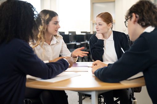 Waist-up view of 15 and 16 year old classmates in school uniforms sitting at round table in collaborative work environment and debating educational subjects.